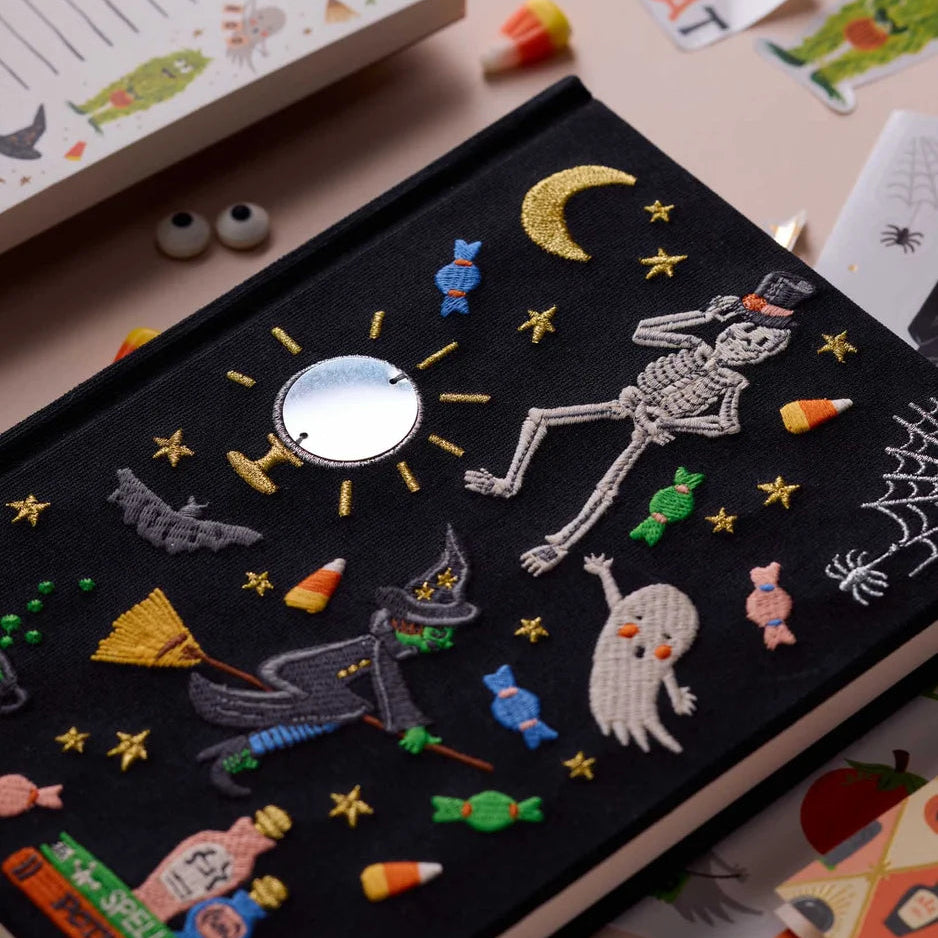 A close-up of a black fabric-covered  journal decorated with embroidered Halloween motifs, including a skeleton, moon, stars, candy, a witch riding a broomstick, a ghost, bats, and cobblestones. A small round mirror is stitched in the center.