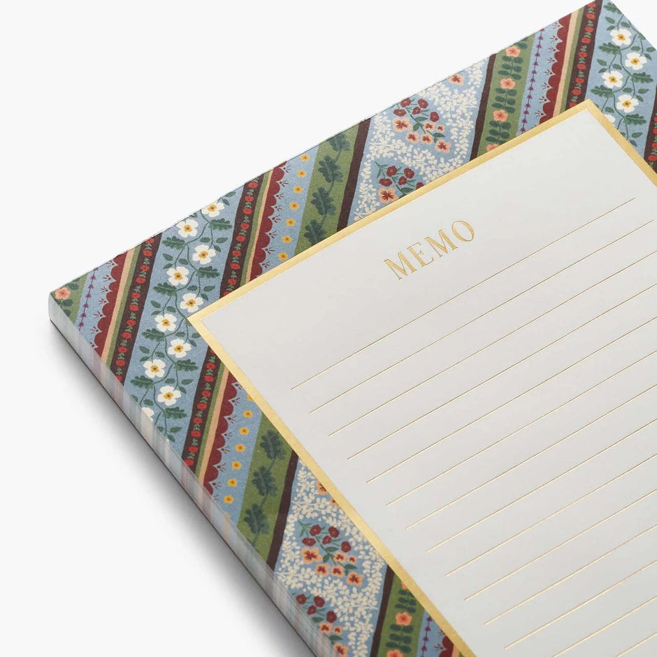 A close-up of a memo pad with a cream-colored lined writing area and the word 'MEMO' printed in gold, bordered by a colorful patterned cover featuring floral and striped motifs in blue, green, red, and gold.