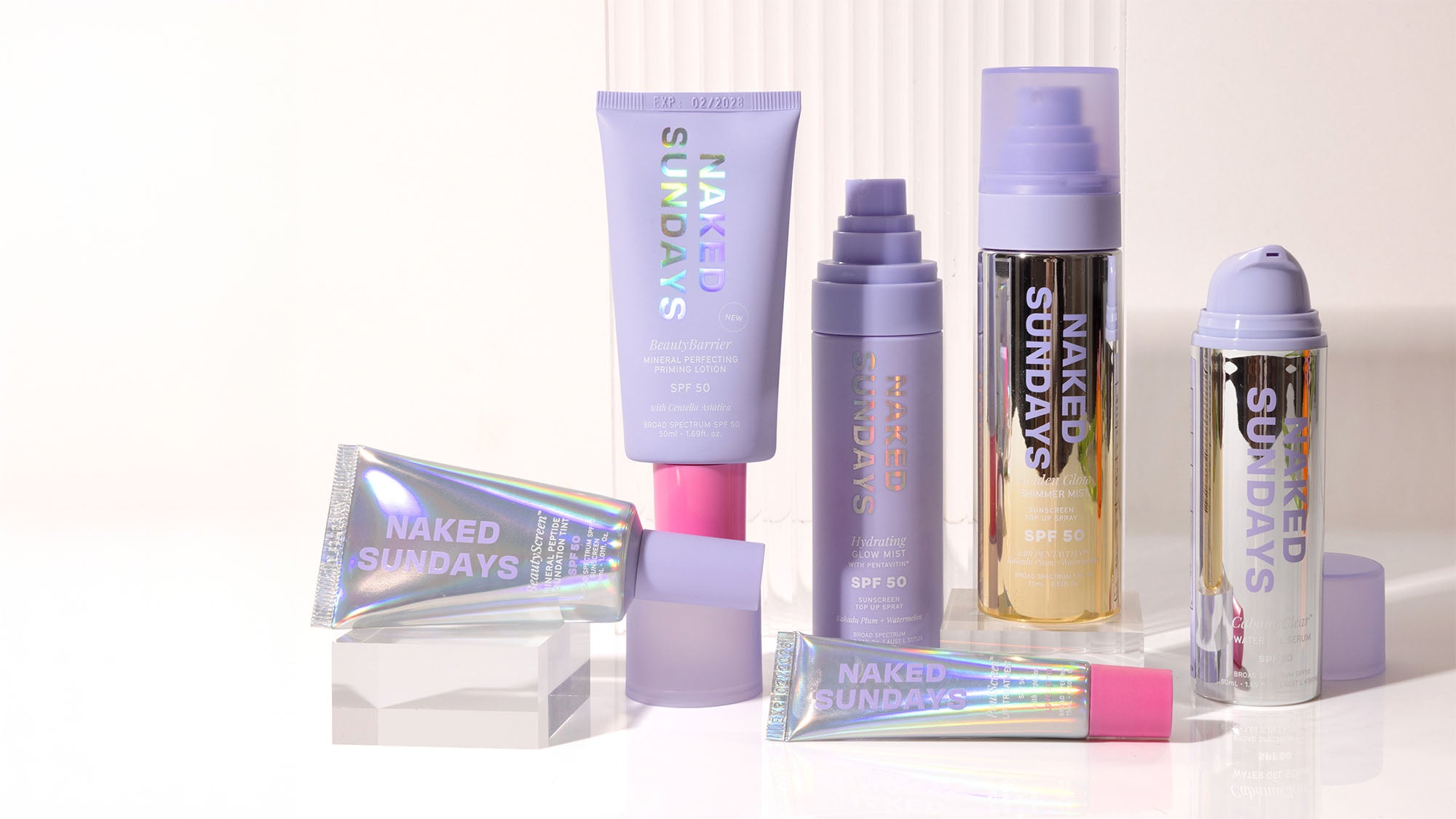 A group of NAKED SUNDAYS skincare and makeup products arranged on a clean white studio background. The set includes a lavender tube, a purple spray bottle, a gold-and-lavender bottle, a silver aerosol can, a holographic tube with a pink cap, and a small clear block prop.