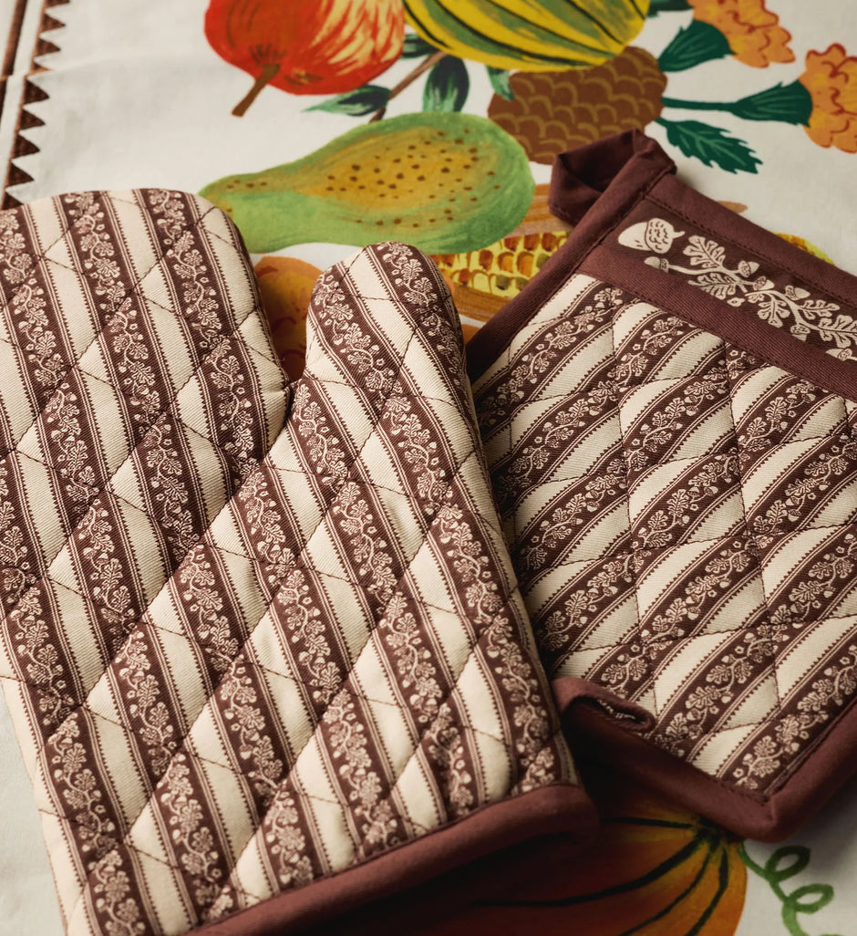 Two quilted oven mitts with brown and cream floral striped patterns, displayed on a colorful fruit-patterned tablecloth.