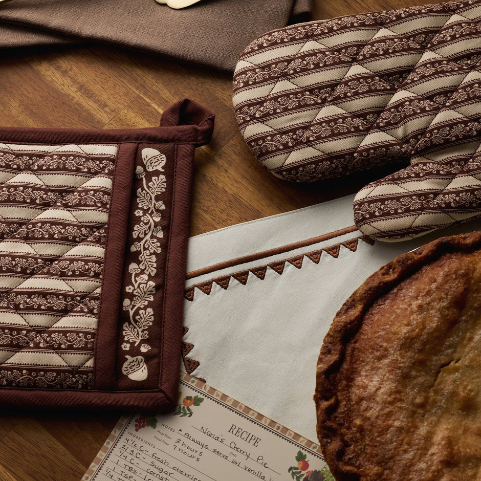 A quilted fabric oven mitt and matching pot holder with brown and beige striped and floral patterns, styled on a wooden table alongside a pie, recipe card, and other kitchen textiles.