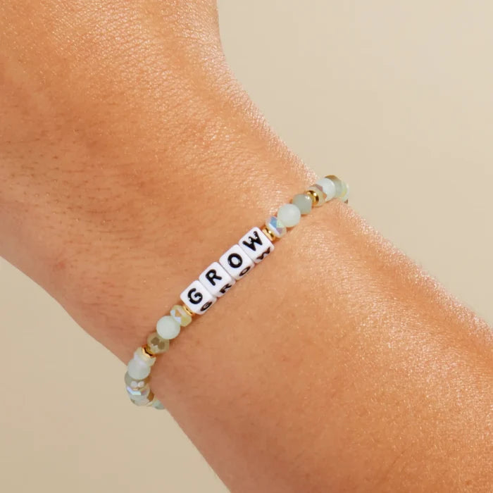 Grow, A close-up of a person's wrist wearing a beaded stretch bracelet with white letter beads spelling 'GROW' and light blue-green faceted beads separated by small gold spacer beads, against a warm beige background.