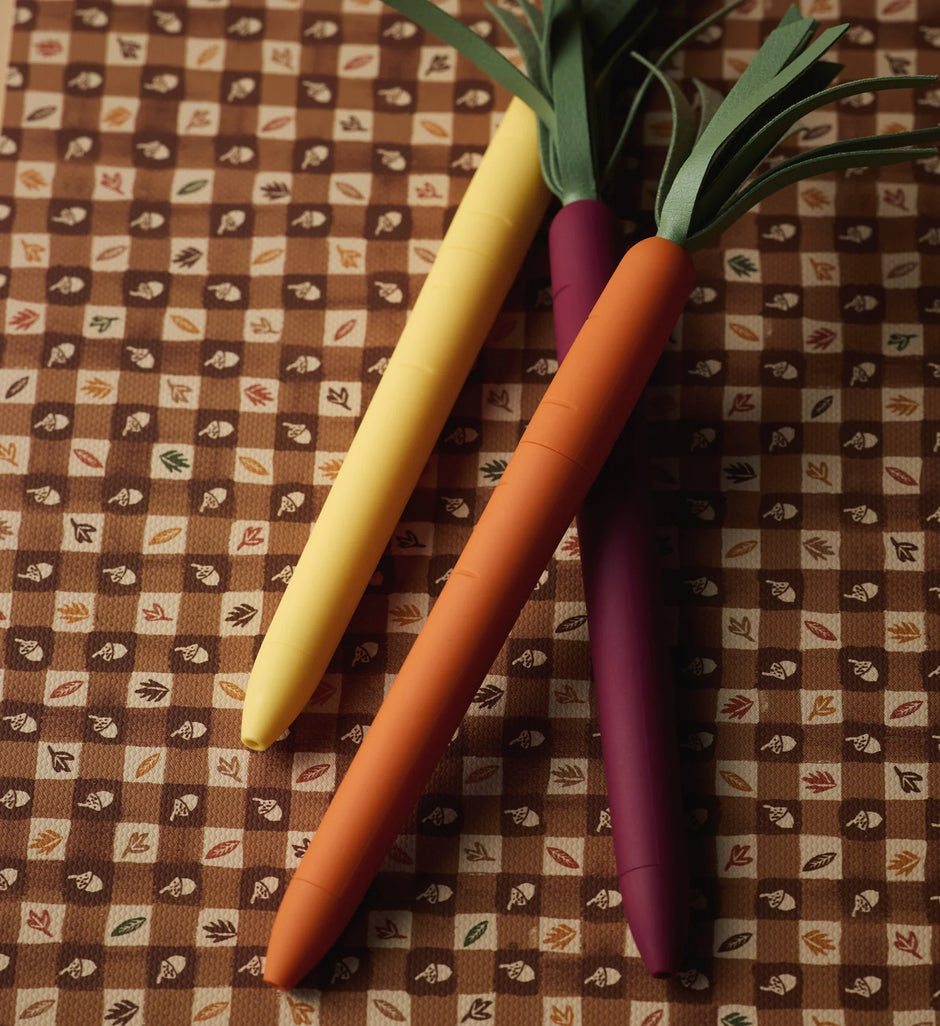 Three carrot-shaped pens with green leaf tops are arranged diagonally on a brown and beige patterned fabric background. The pens are colored yellow, orange, and purple.