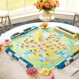 A colorful mahjong game set displayed on a round white table in a bright room. The board features a pastel illustration with cranes and birds, surrounded by blue racks holding tiles, with small bowls, a vase of flowers, and tea cups nearby.
