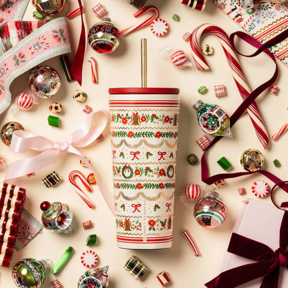 A festive holiday-themed tumbler with a gold metal straw, decorated with a red and green Christmas pattern, surrounded by candy canes, ornaments, ribbons, and wrapped gifts on a light cream background.
