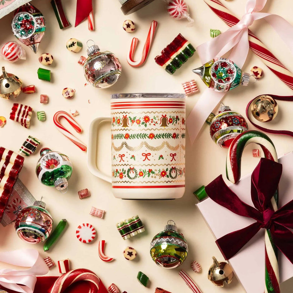A festive Christmas-themed mug with a red and green holiday pattern, surrounded by candy canes, ornaments, ribbons, and wrapped gifts on a light cream background.