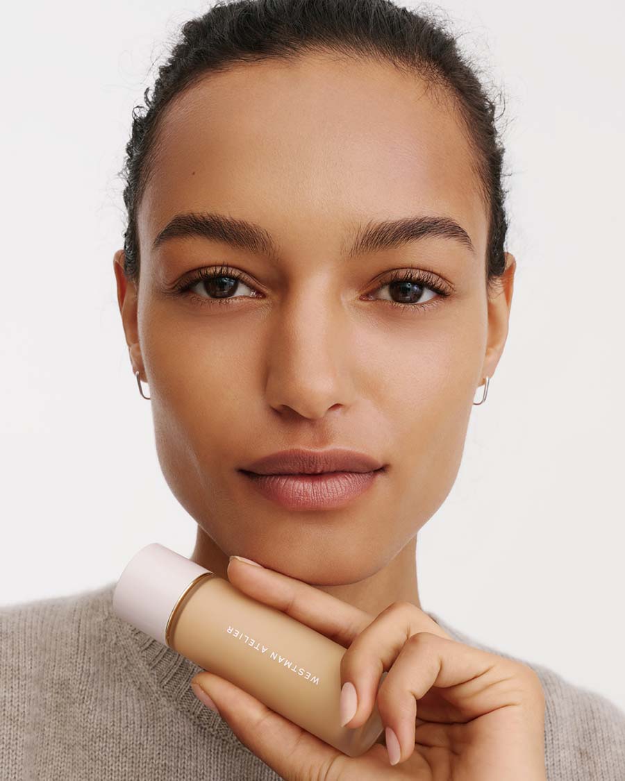 A close-up of a woman holding a bottle of Westman Atelier liquid foundation against a clean white background.