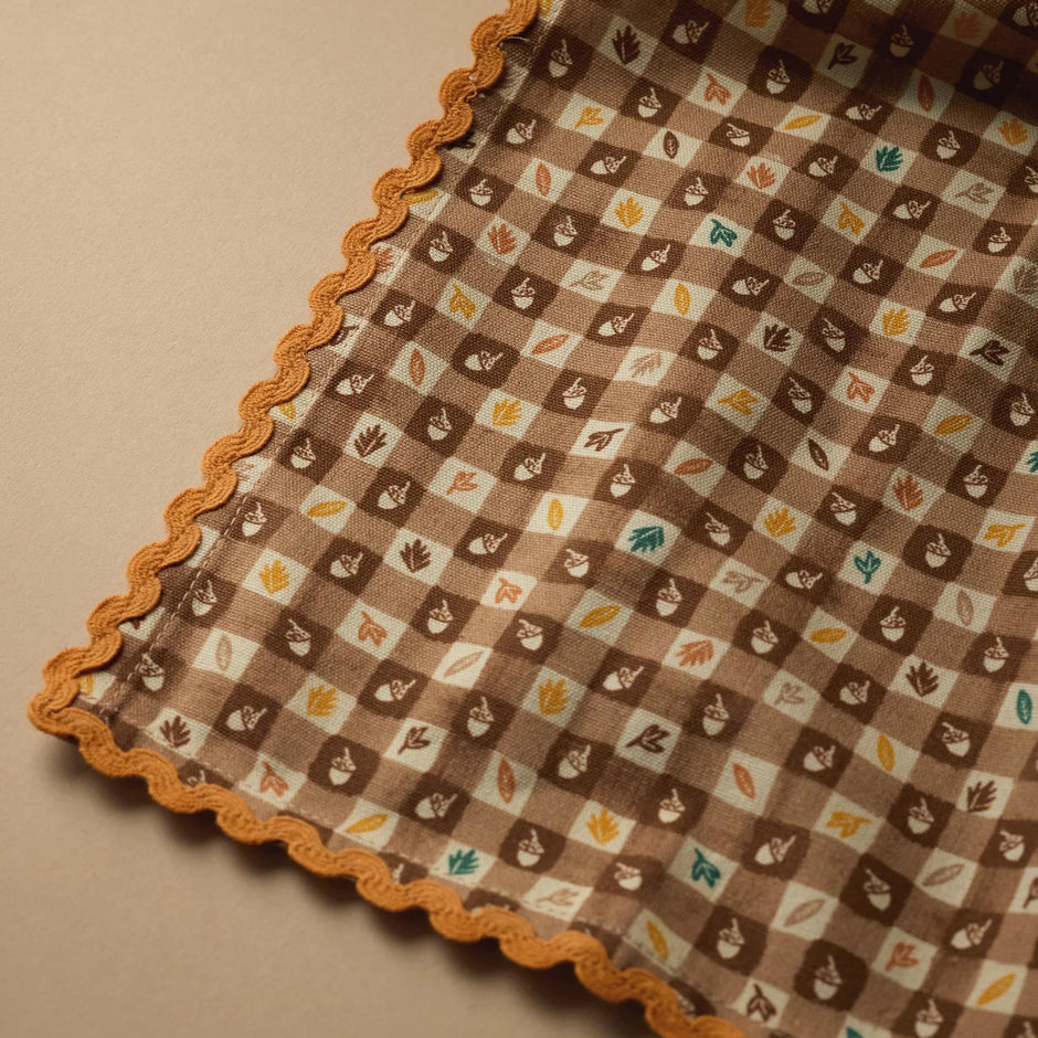 A close-up of a patterned fabric tea towel with a scalloped orange border, featuring a brown and white gingham check with small leaf motifs, on a beige background.