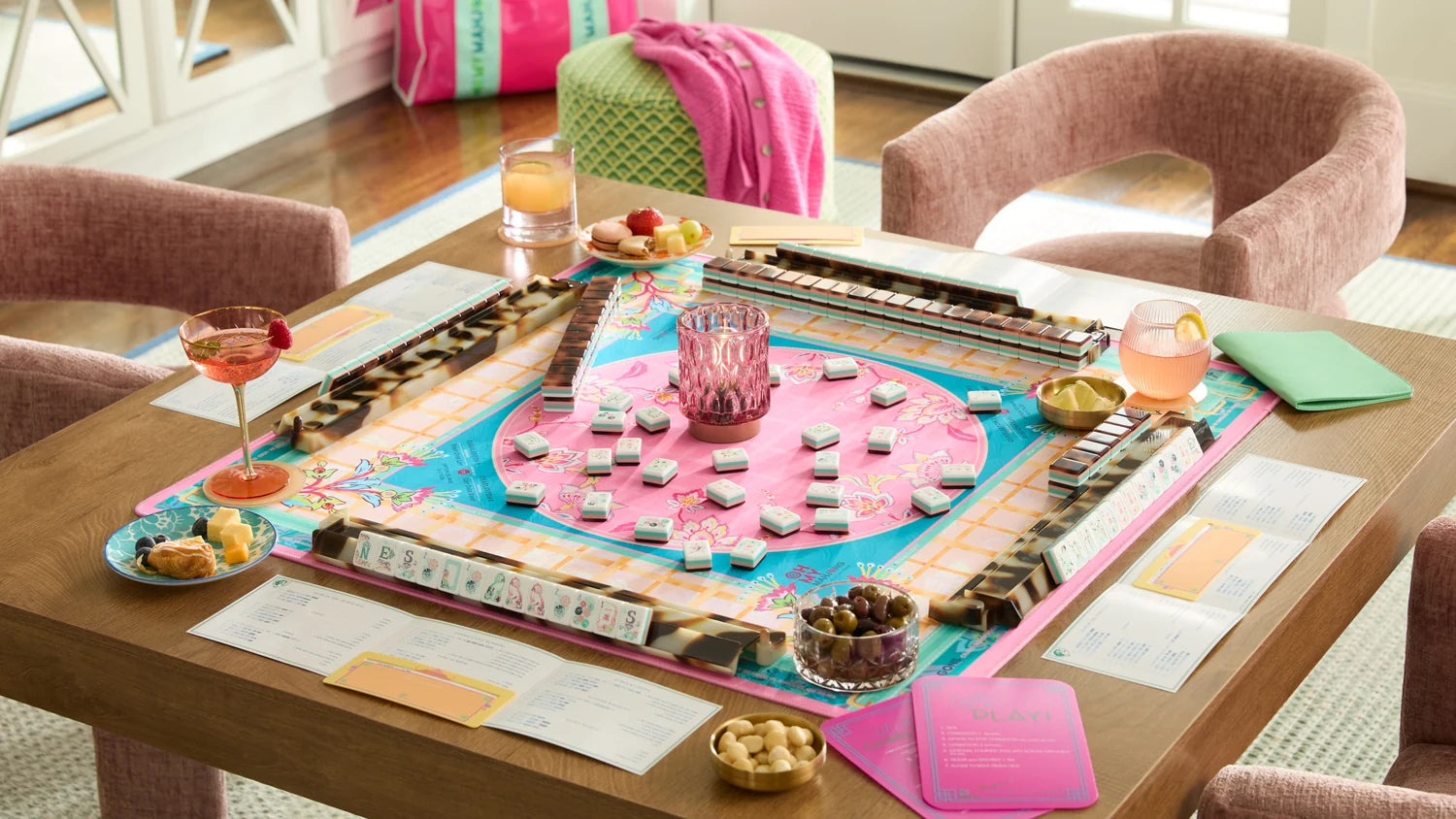 A colorful Mahjong game set up on a wooden dining table surrounded by pink upholstered chairs, with drinks, snacks, and game accessories arranged on the tabletop in a bright indoor room.