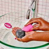 Person holding a pink and black scrubber over a sink with soap suds.