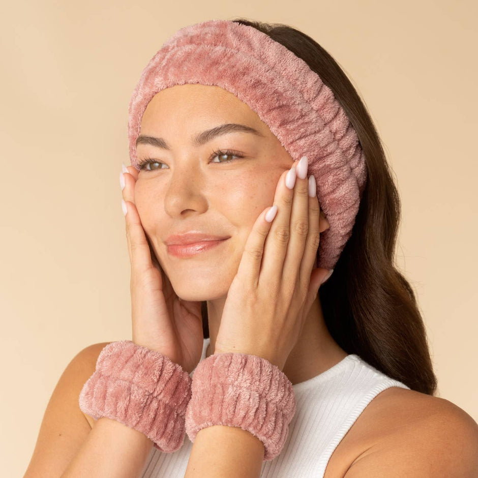 Woman wearing pink spa headband and wristlets set for skincare routine on neutral background