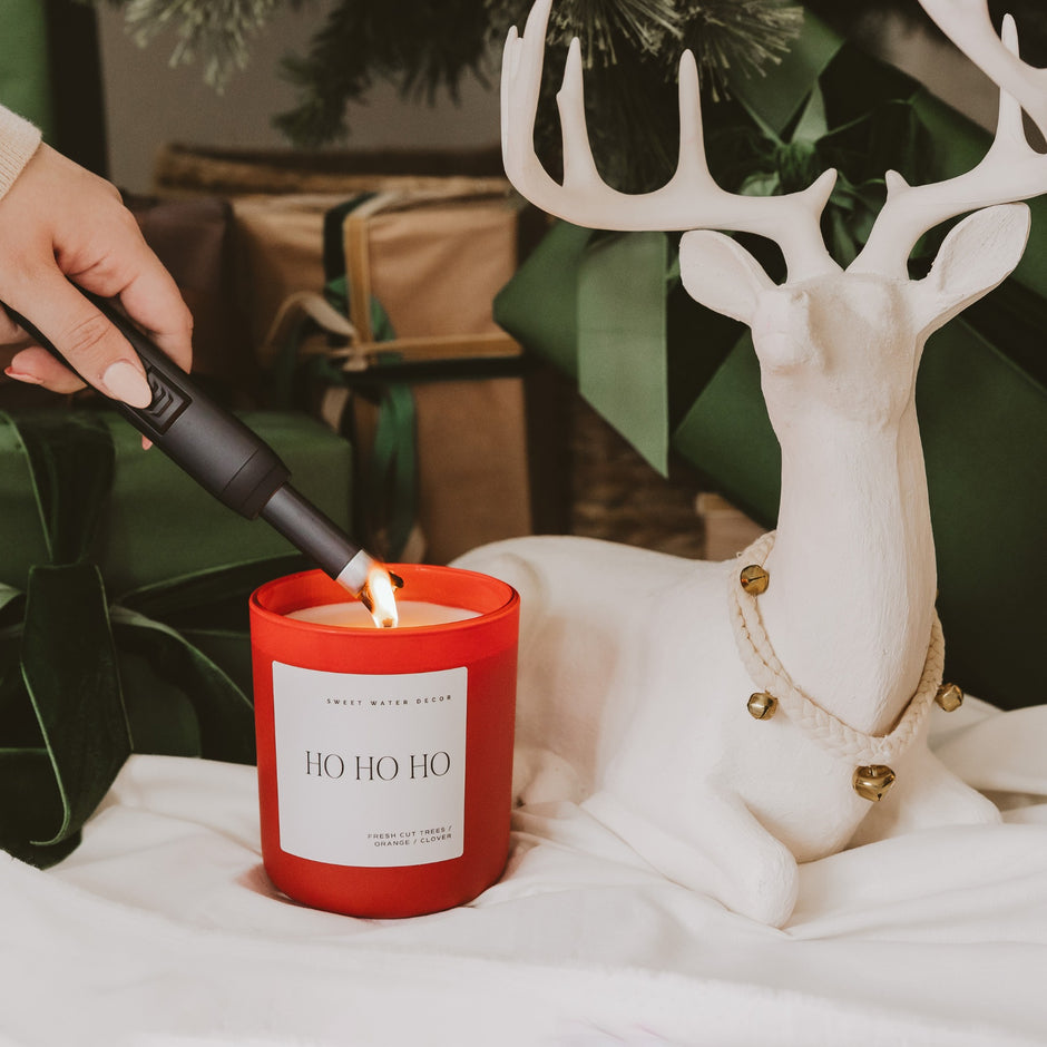 Ho Ho Ho red soy candle being lit in festive holiday setting with white deer figurine and greenery
