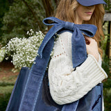 Woman wearing denim bow tote bag with white cable knit sweater, holding baby's breath flowers in garden setting