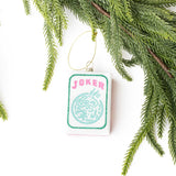 Joker mahjong tile glass ornament hanging from gold string among evergreen branches