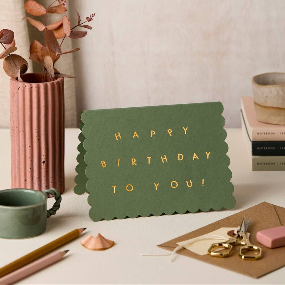 Green scallop edge birthday card with gold text displayed in styled desk setting with flowers and stationery