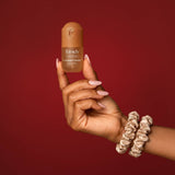 Hand holding Kitsch Caramel Cream hair perfume bottle against red background with accessories