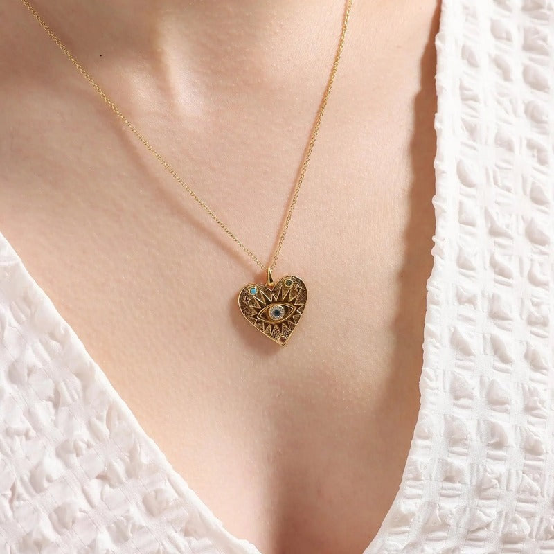 Gold heart evil eye necklace worn on model's chest, featuring ornate design with blue stone detail