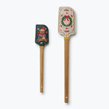Two wooden spatulas with holiday designs: one with a Christmas bear on dark green, one with a wreath on striped cream background.