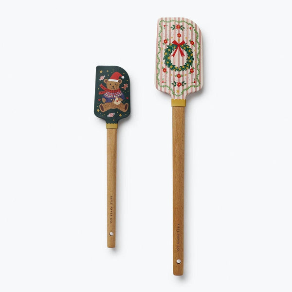 Two wooden spatulas with holiday designs: one with a Christmas bear on dark green, one with a wreath on striped cream background.