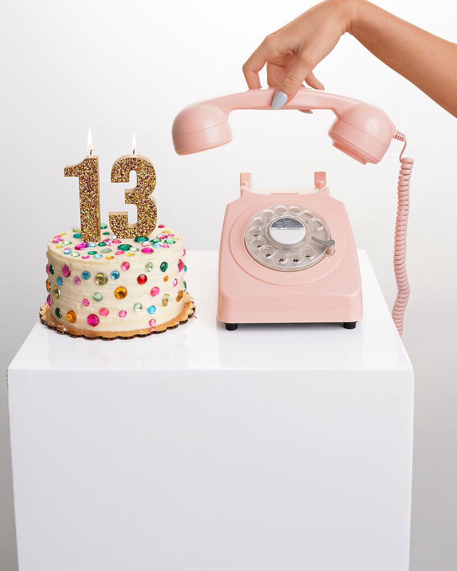A pink retro rotary telephone with a coiled cord sits on a white pedestal next to a small decorated birthday cake with gold '13' candles. A hand is holding the receiver of the phone against a clean white background.