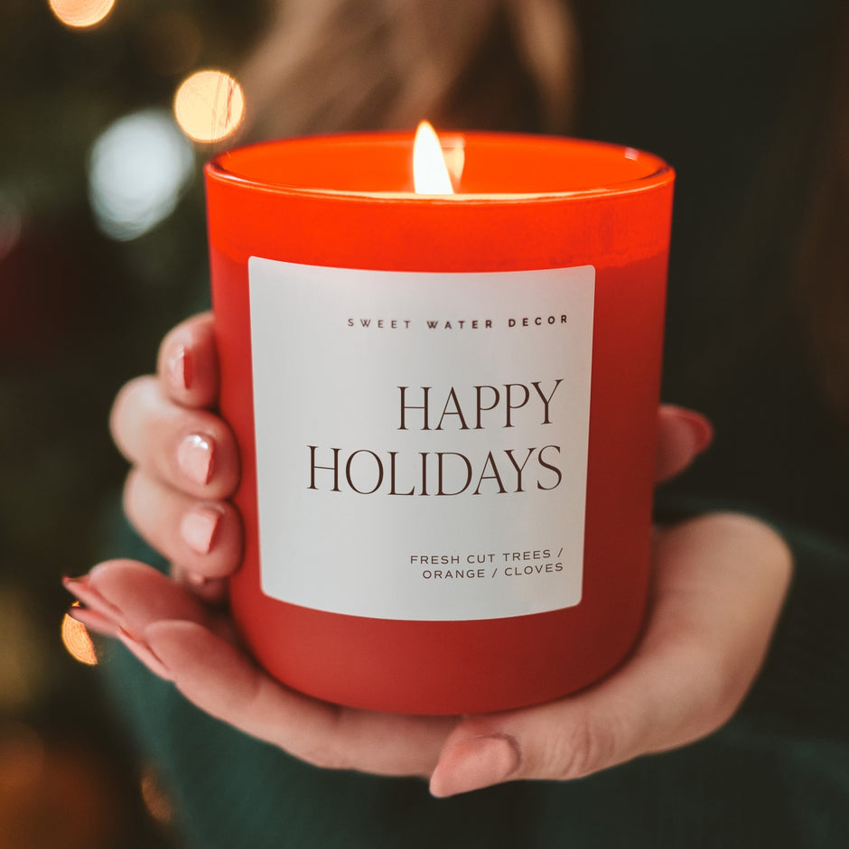 Happy Holidays soy candle by Sweet Water Decor held in hands with warm bokeh background