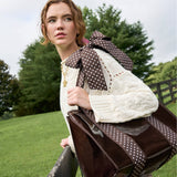 Woman wearing cream cable knit sweater holding brown polka dot bow tote bag outdoors in green field