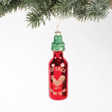 Red hot sauce bottle ornament with green glitter top hanging from evergreen branch