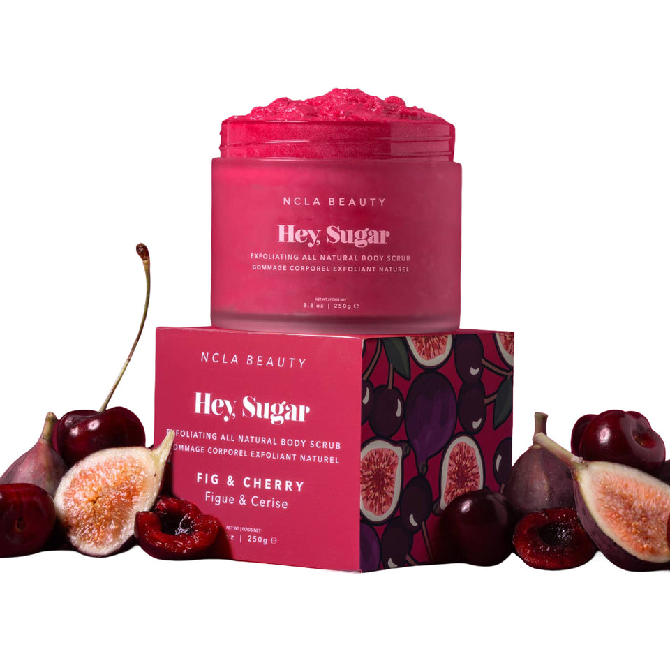 NCLA BEAUTY Hey Sugar Fig & Cherry exfoliating body scrub in a pink jar, displayed on top of its matching pink box with fruit illustrations, surrounded by cherries and sliced figs on a white background.