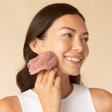 Woman holding pink microfiber face towel against cheek, smiling against neutral background