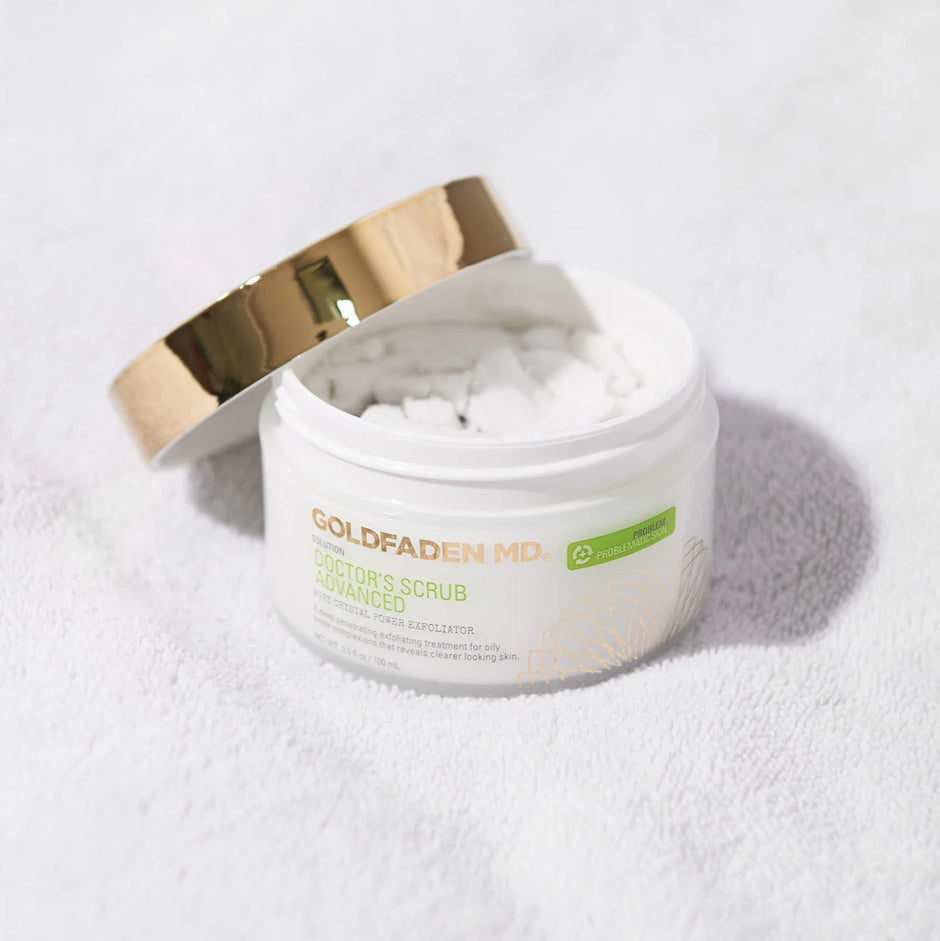 An open jar of Goldfaden MD Doctor's Scrub Advanced with its gold lid resting beside it, set on a white towel.