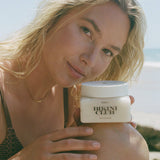 Woman holding a jar of 'Bikini Club' product on a beach.