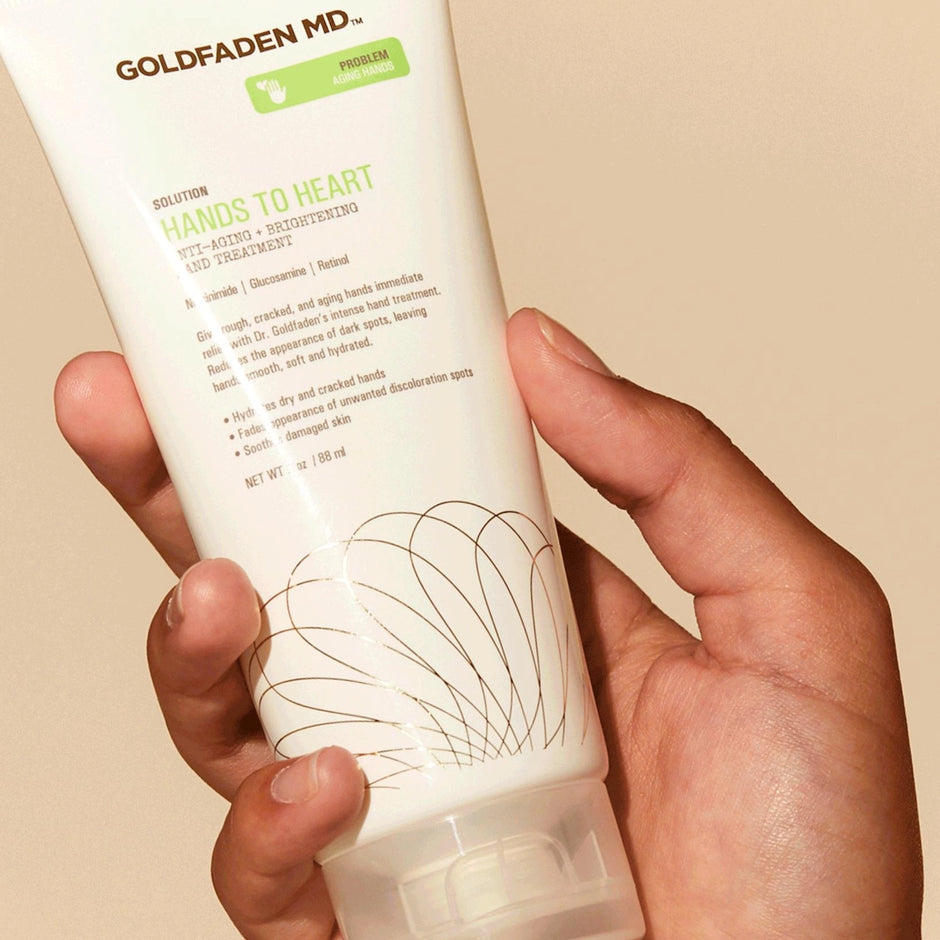 A hand holding a tube of Goldfaden MD Hands to Heart Anti-Aging + Brightening Hand Treatment.