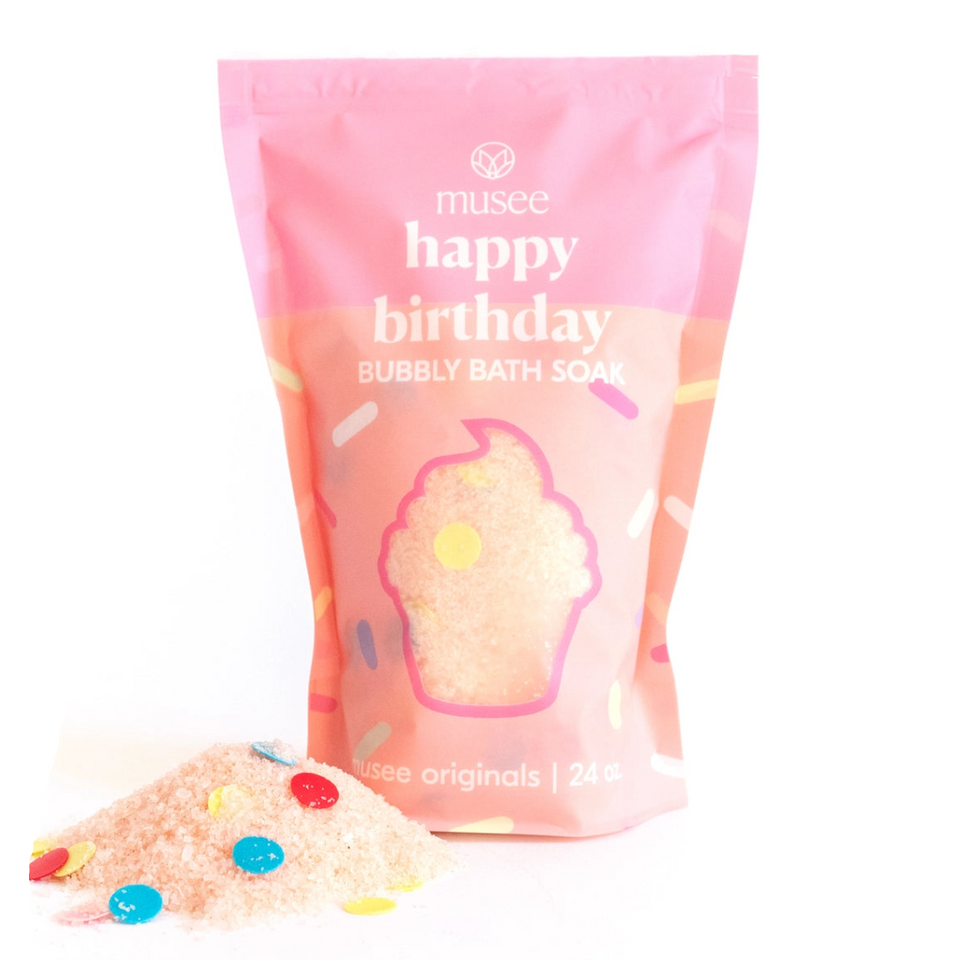 A pink stand-up pouch of musee 'happy birthday' Bubbly Bath Soak with a cupcake graphic and colorful confetti, shown on a white background with some bath soak spilled in front.