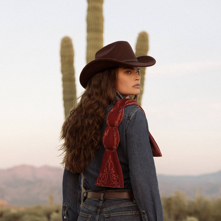 Woman wearing cowboy hat and denim shirt with red bandana, standing in desert landscape with saguaro cactus
