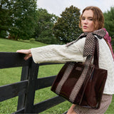 Woman wearing cream cable knit sweater holding brown polka dot bow tote bag by fence in pastoral setting