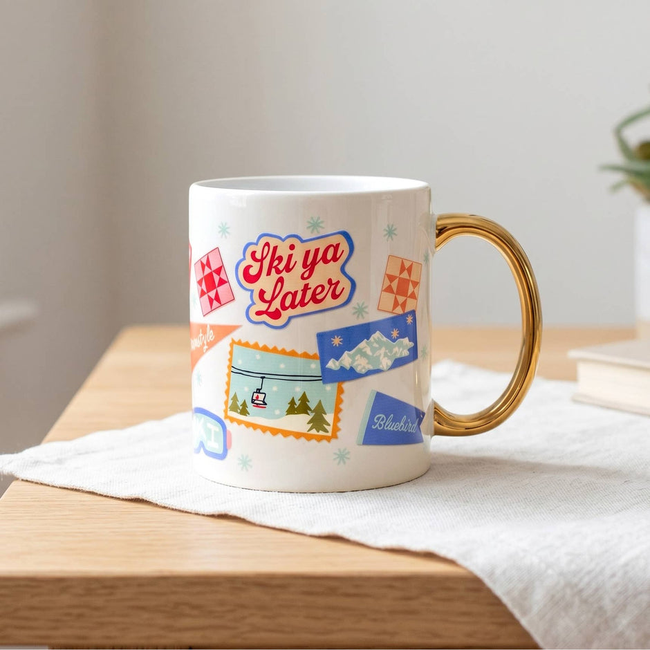 Ski Ya Later ceramic mug with gold handle on wooden table with white cloth