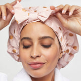 Woman wearing blush pink Kitsch shower cap with gold polka dot pattern, adjusting the bow