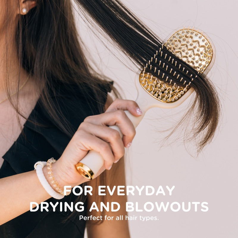 Woman holding tan vented hair brush while styling long dark hair, wearing gold rings and bracelets