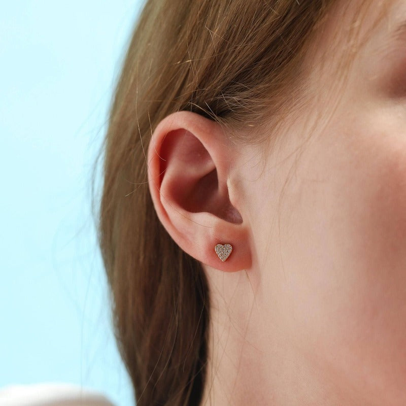Gold heart stud earrings with CZ accents worn on model's ear against blue background