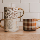 Three decorative ceramic mugs with floral and plaid patterns displayed on wooden surface