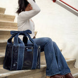 Woman sitting on stairs wearing white sweater and jeans, holding denim bow jelly tote bag