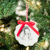 White scalloped photo ornament with red ribbon bow hanging on decorated Christmas tree with lights