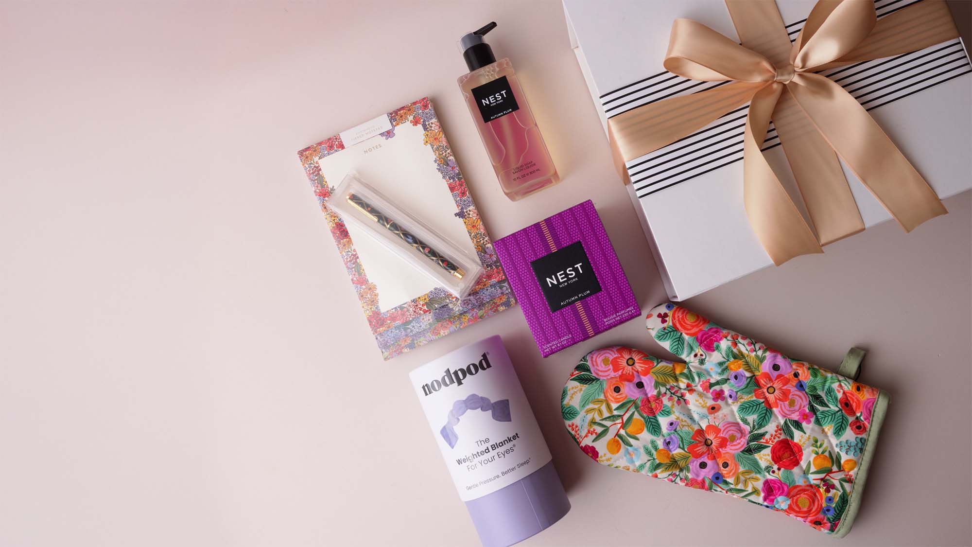 A curated gift collection featuring a white gift box with a gold ribbon, a pink hand wash bottle, a floral notepad and pen, a purple box, a nodpod, and a floral oven mitt, arranged on a light pink background.