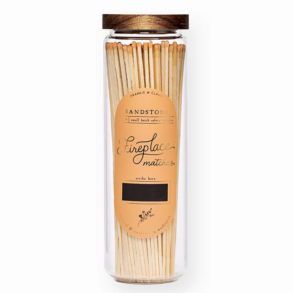 Sandstone, tall glass apothecary jar of burnt orange tipped fireplace matches with acacia wood lid and gold foil label on a white background