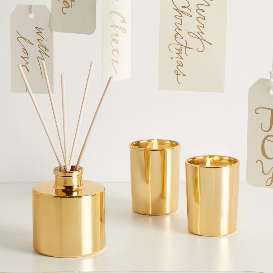A gold-toned reed diffuser bottle with several reeds inserted, styled alongside two matching gold votive candle holders on a white surface against a white background. Hanging paper tags with holiday greetings are partially visible behind the items.