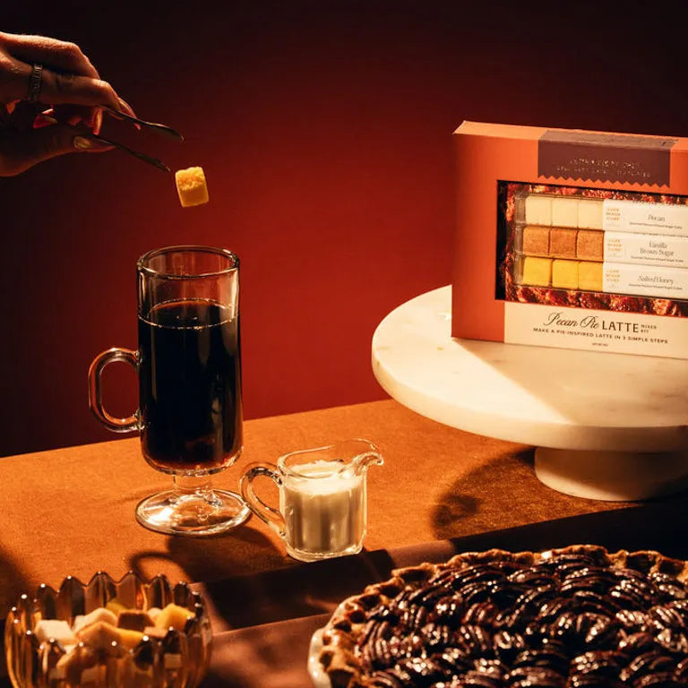 Hand dropping a cube into a glass mug of coffee beside the Pecan Pie Latte kit