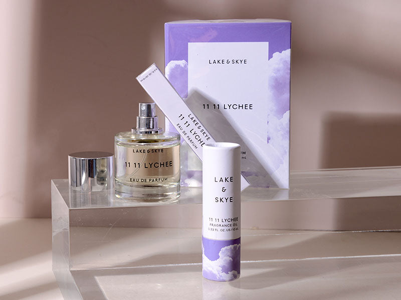 Lake & Skye 1111 Lychee fragrance set on a clear acrylic riser, featuring a glass perfume bottle, a cylindrical tube, a box, and a matching applicator wand against a warm beige background.