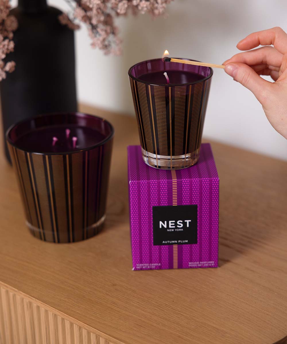 A Nest Autumn Plum scented candle in a dark purple and gold striped glass jar, being lit with a wooden match by a hand. The second unlit candle and its purple patterned packaging box sit on a light wood table.