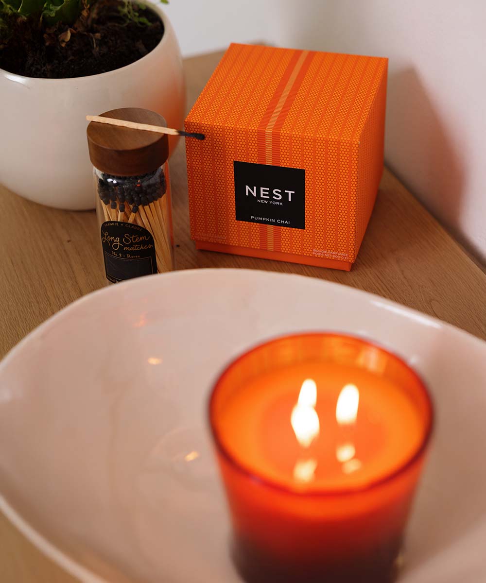 A Nest Pumpkin Chai scented candle in an orange glass holder sits on a white tray, with its orange textured box and a small match container behind it on a wooden table.