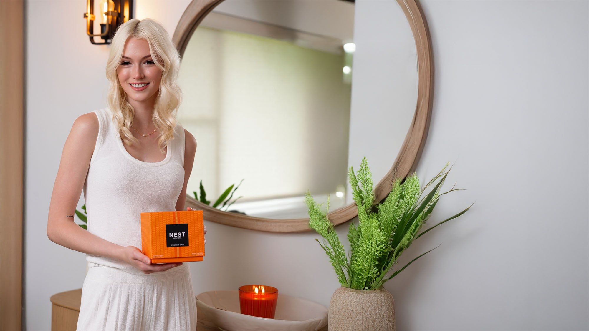A blonde woman in a white sleeveless top stands indoors holding an orange box labeled NEST. The setting includes a large round wooden-framed mirror, a potted green plant, and a lit orange candle on a table.